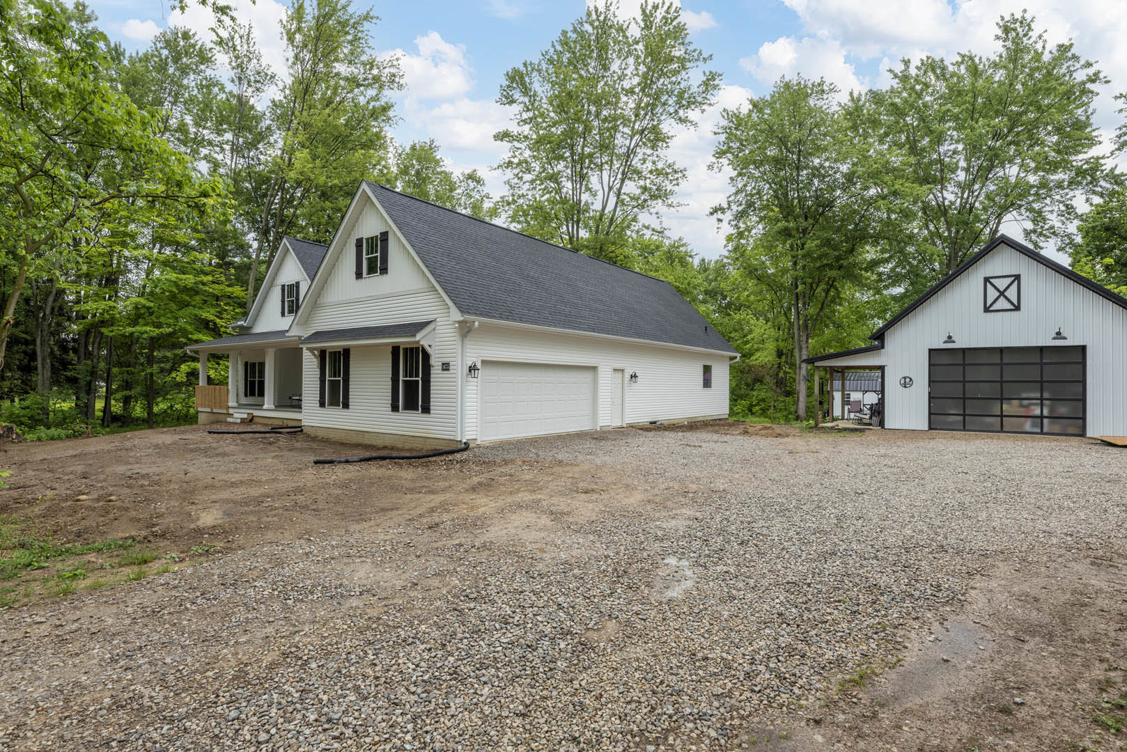 White house with two attached garages, gravel driveway, black-framed windows, gabled roof, mature trees in background, partly cloudy sky