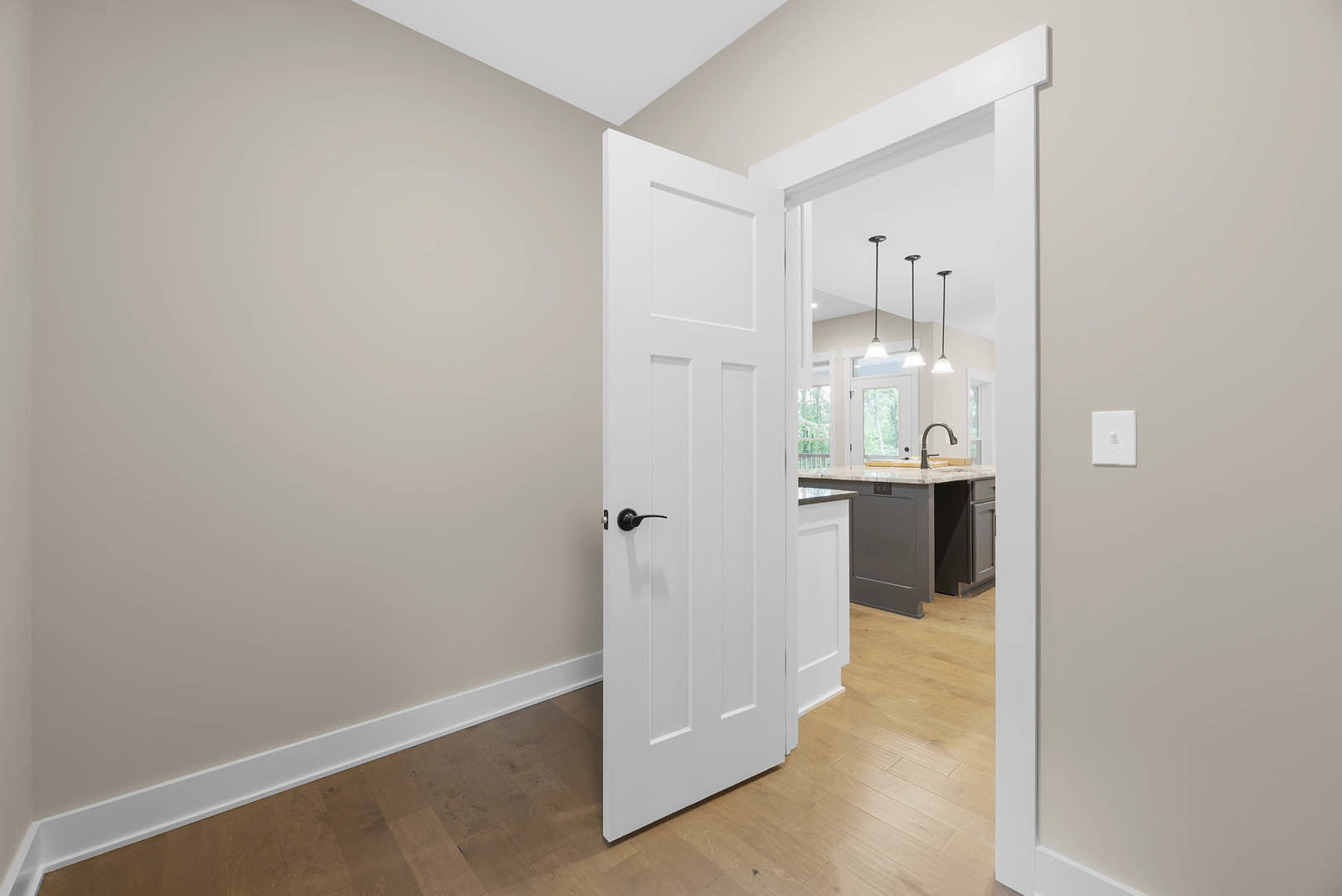 White paneled door with brushed metal handle, white light switch on plaster wall, laminate flooring, partial view of kitchen counter with chrome faucet, overhead pendant lights