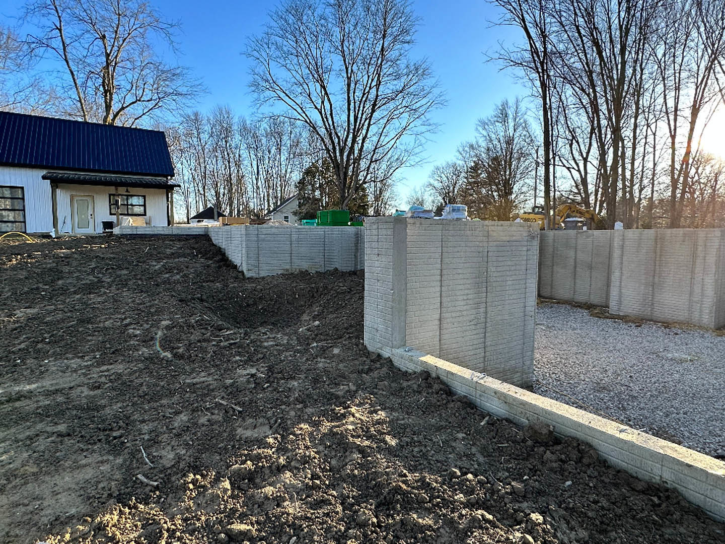 White house with blue roof behind a concrete wall, leafless tree in foreground, dirt ground and sparse plants adjacent to wall, black-barred window visible.