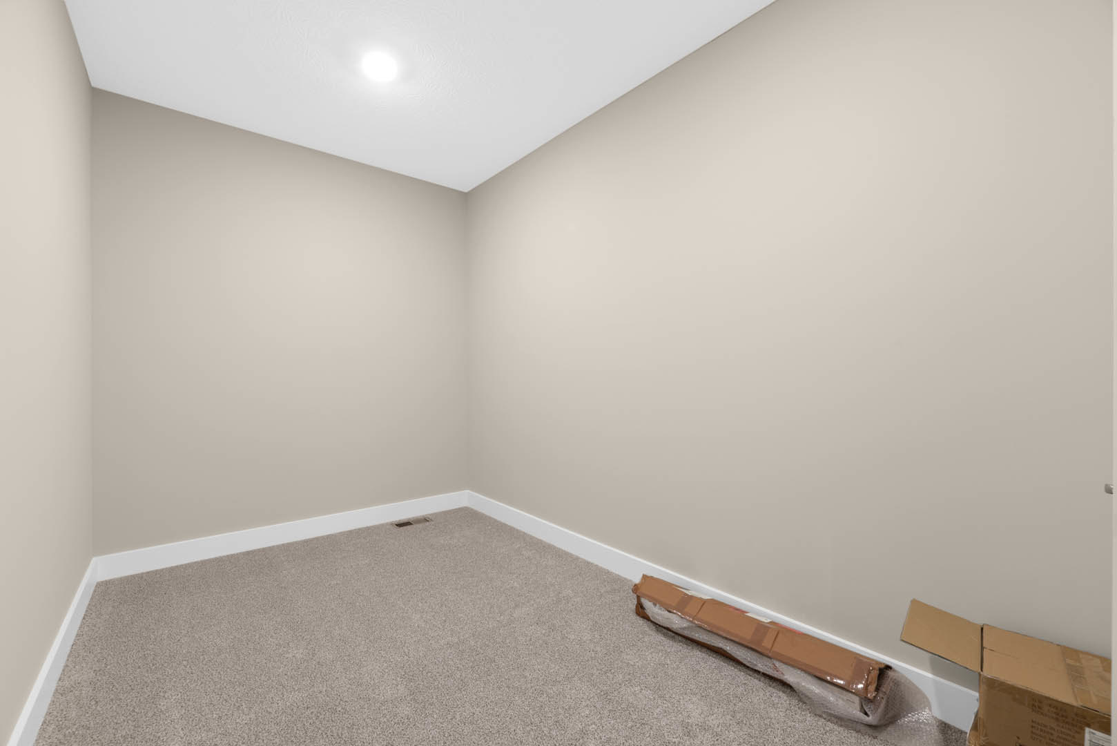 Carpeted room corner with a cardboard box wrapped in plastic, white walls, ceiling light, and a wooden shelf