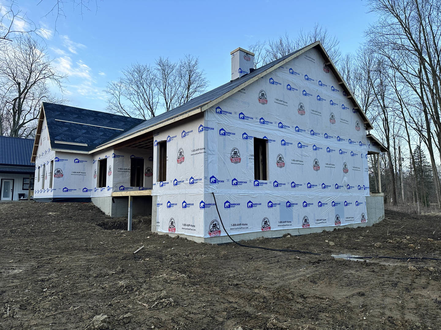 Partially built house with white exterior walls, black hose attached, blue tarp covering roof, plastic sheeting, exposed chimney, and patch of dirt in foreground