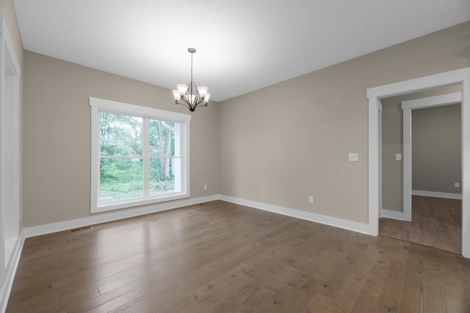 Spacious room with hardwood flooring, ornate chandelier, large window overlooking trees, white door with matching trim, plaster walls, and decorative molding