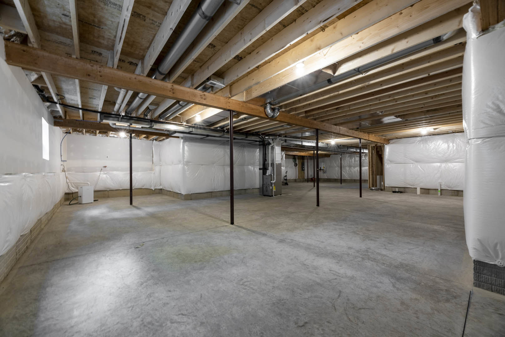White-walled basement with exposed wooden ceiling beams, visible pipes, concrete floor, steel support pole, plastic wall covering, and stacked white bags.