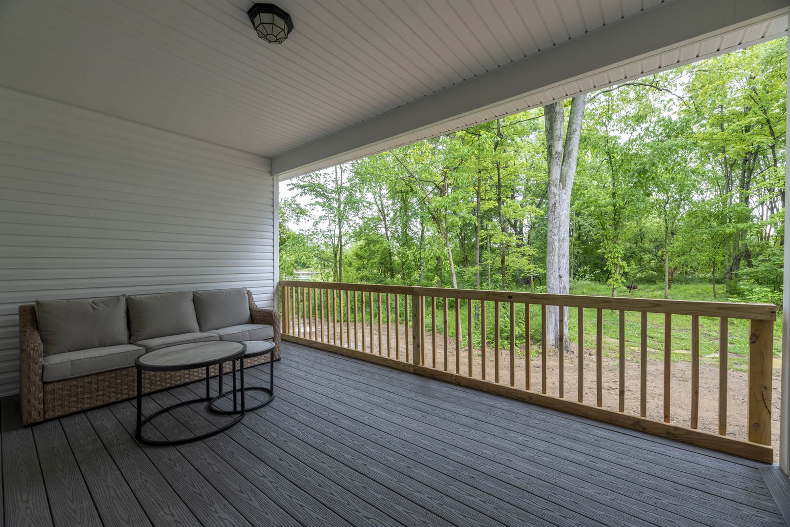 Wooden deck with horizontal railing, outdoor couch and coffee table, lamp on table, surrounded by trees and greenery