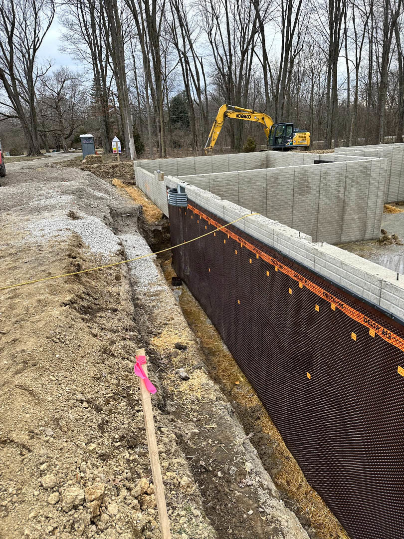 Framed custom home under construction with exposed brick wall, yellow caution tape along dirt ground, wooden stakes, and yellow construction equipment on site
