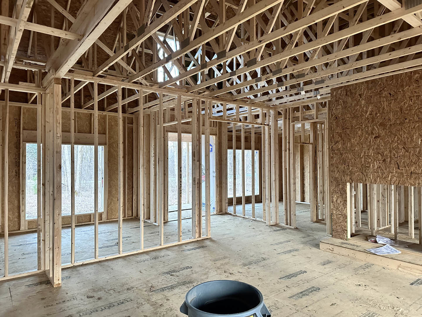 Wood-framed interior under construction with exposed beams, unfinished walls, and a black bucket on the plywood floor