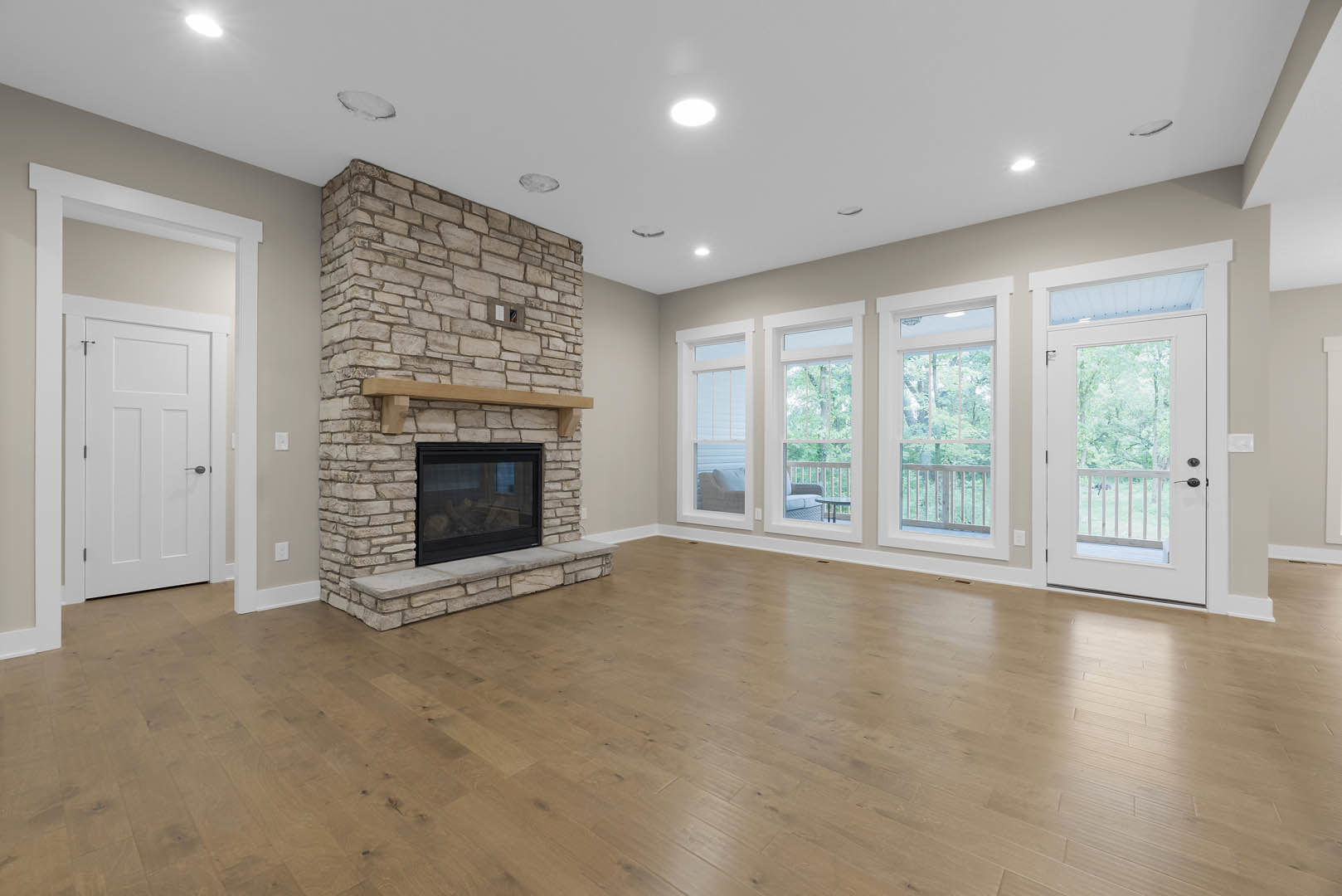 Living room with hardwood flooring, brick fireplace featuring a glass door, exposed wood beam, white door with black handle, and plaster walls.