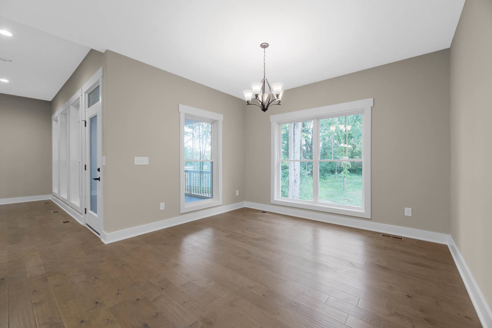 Chandelier hanging from white ceiling in spacious room with hardwood floors, large windows overlooking trees and balcony, plaster walls, and wood trim