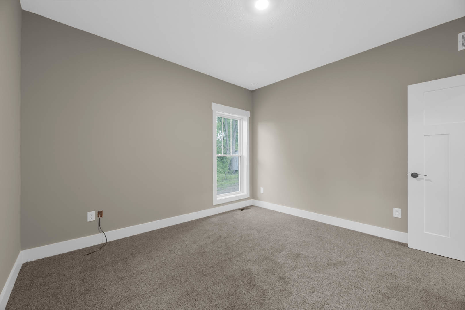 Carpeted room with white walls, large window showing trees outside, white door with black handle, ceiling light fixture, floor vent, and black wire on white surface