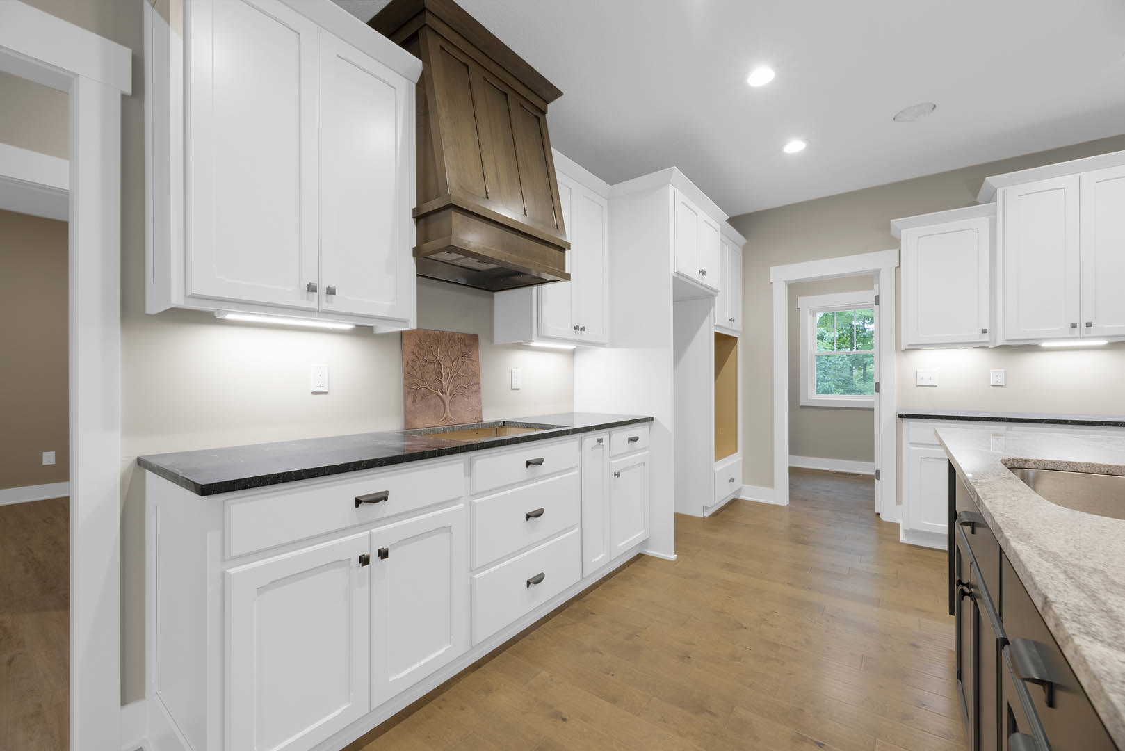 White kitchen with silver-handled cabinets, black countertops, wood flooring, and a tree carving detail on the wall; window reveals leafy trees outside.