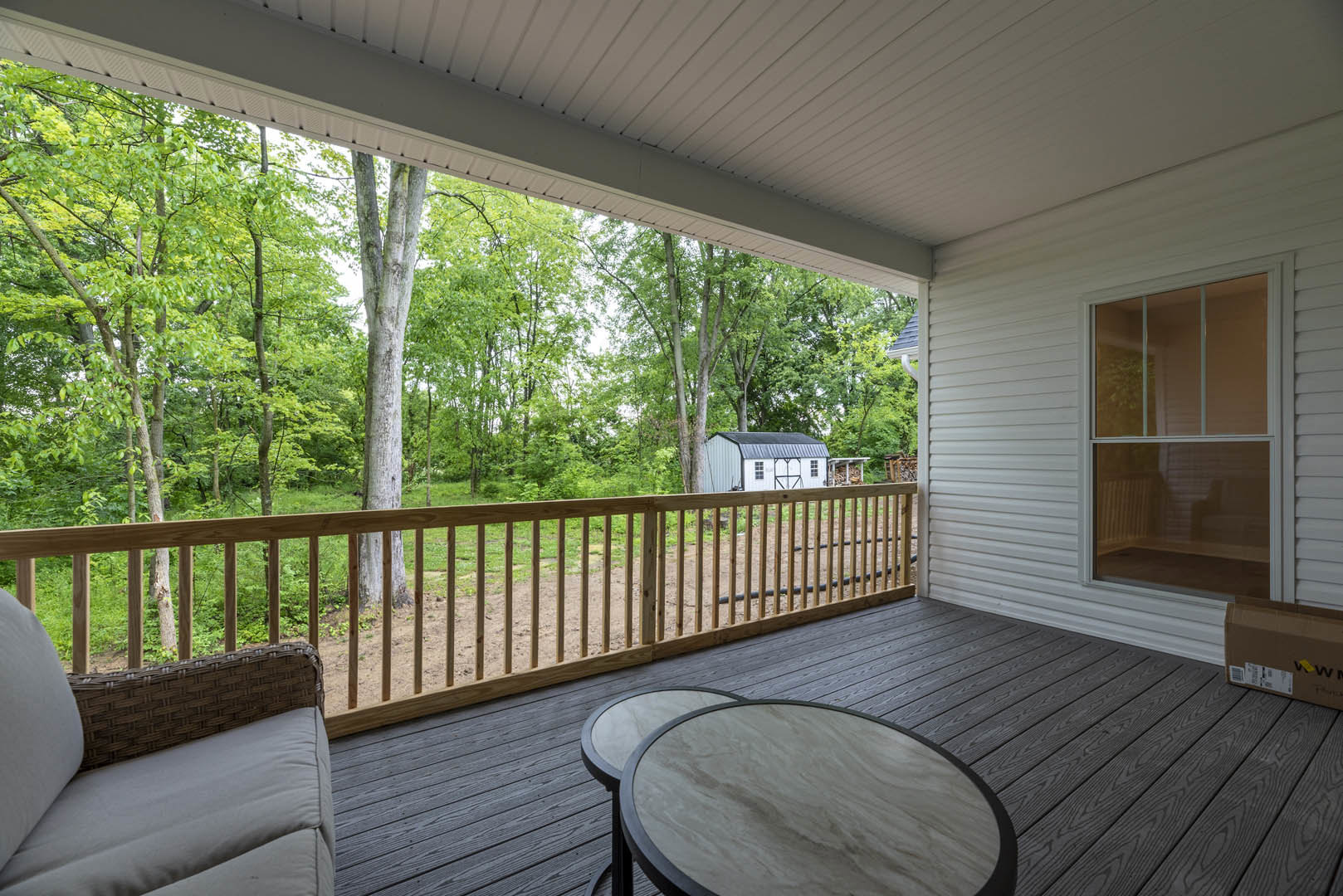 Wooden deck with a circular table and chairs, surrounded by a wooden railing, large glass-paned window, and outdoor trees visible in the background
