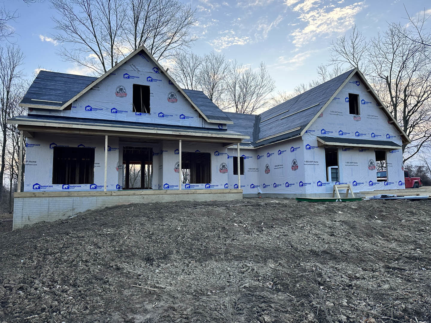 Framed house under construction with blue roof, exposed wood, dirt field in foreground, mature trees and cloudy sky in background