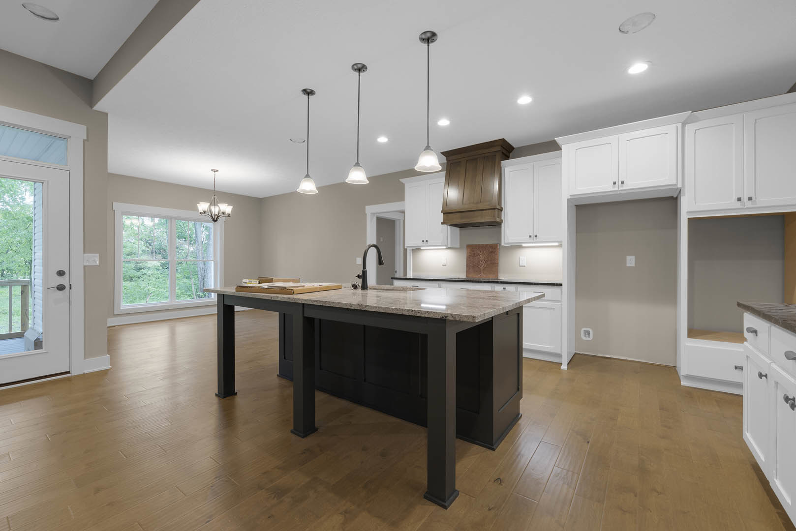 Kitchen with wood flooring, central island featuring black and white countertop, wooden cabinets, white ceiling, window overlooking trees, white door with lock, and white object on