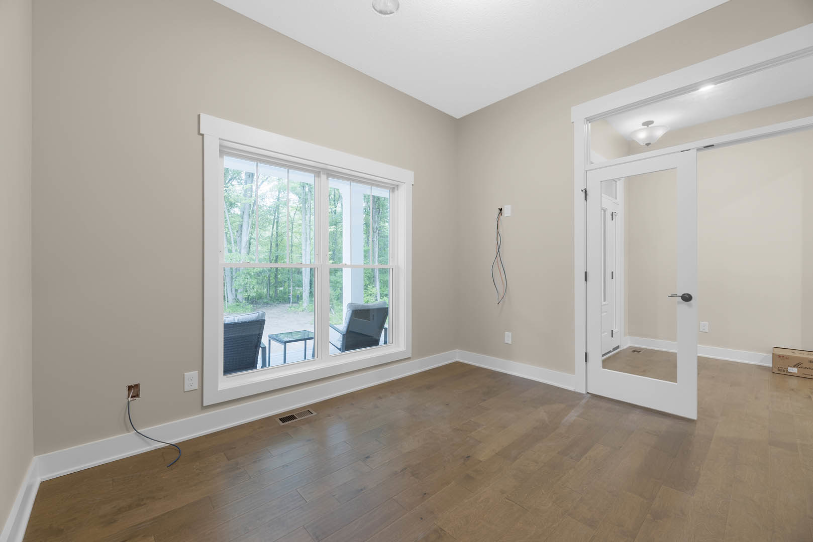 Spacious room with light wood flooring, large wall mirror, glass table with metal legs, upholstered chair, white plaster walls, window overlooking outdoors, decorative molding