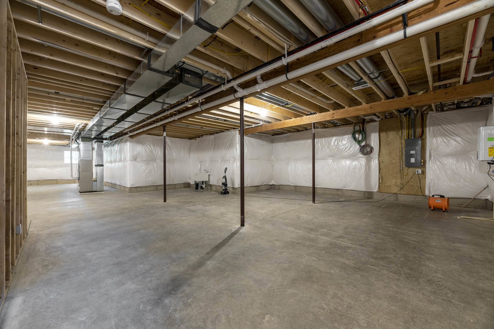 Room under construction with white plastic coverings on walls and floor, exposed wood ceiling beams, visible pipes, orange cylinder with black handle, and concrete floor partially