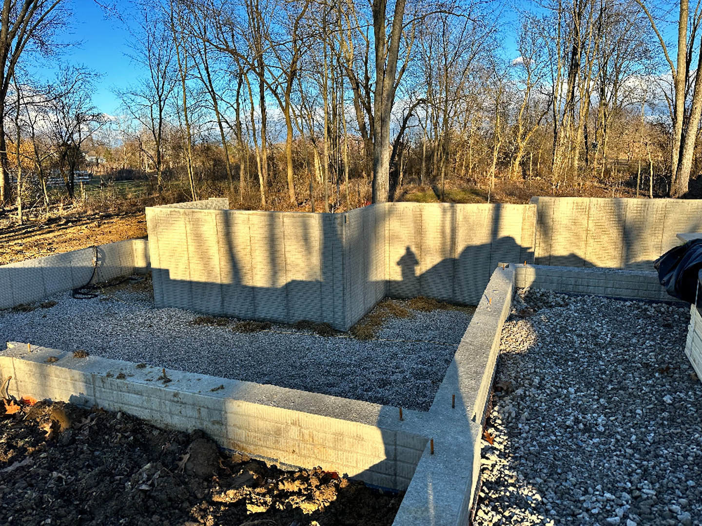 Smooth concrete wall casting a distinct shadow onto gravel and dirt, bordered by a pile of gravel and surrounded by trees and plants under an open sky