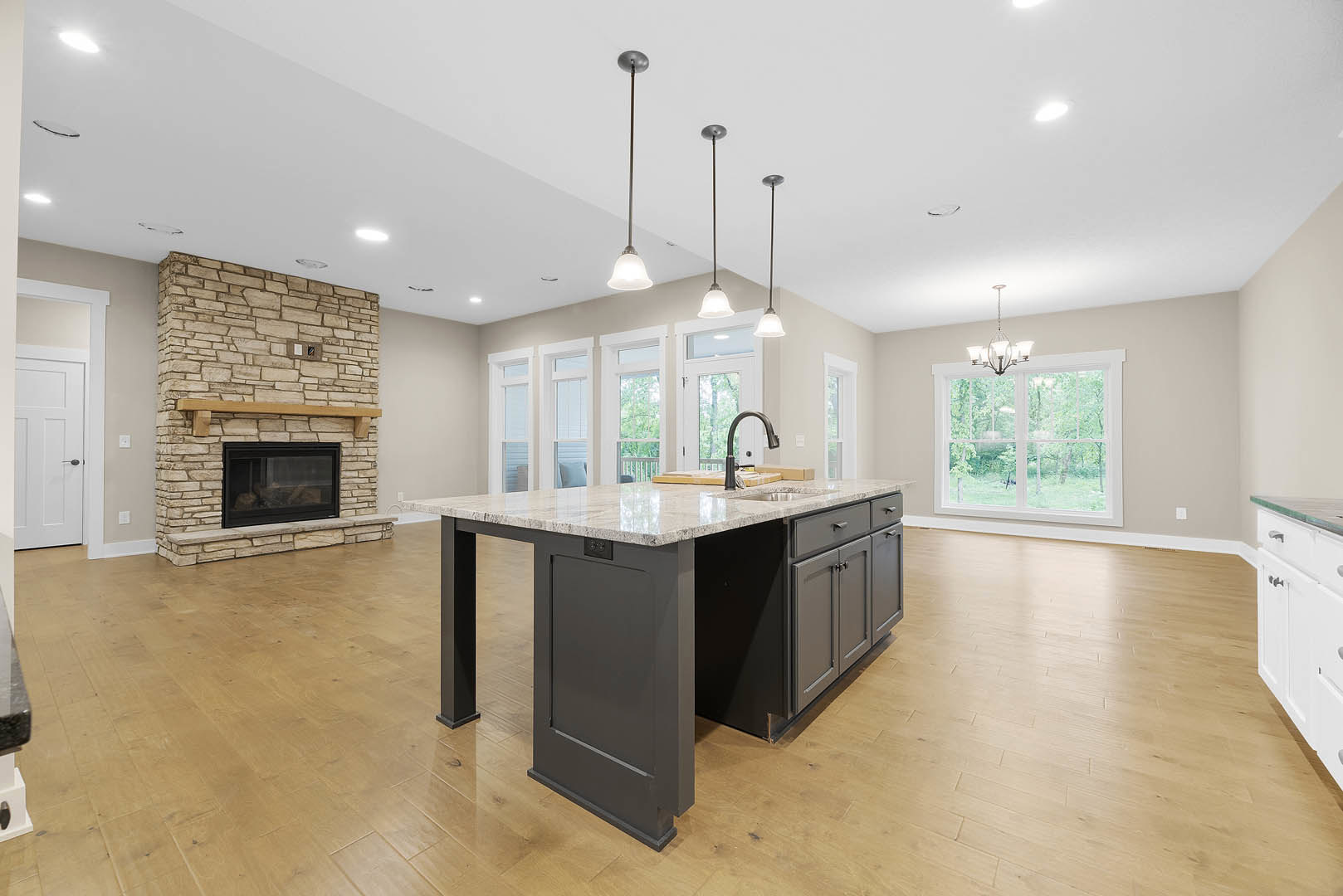 Spacious kitchen featuring a large island with built-in sink, white cabinetry, stone countertops, and a fireplace with a wood mantel and glass window, adjacent to windows offering