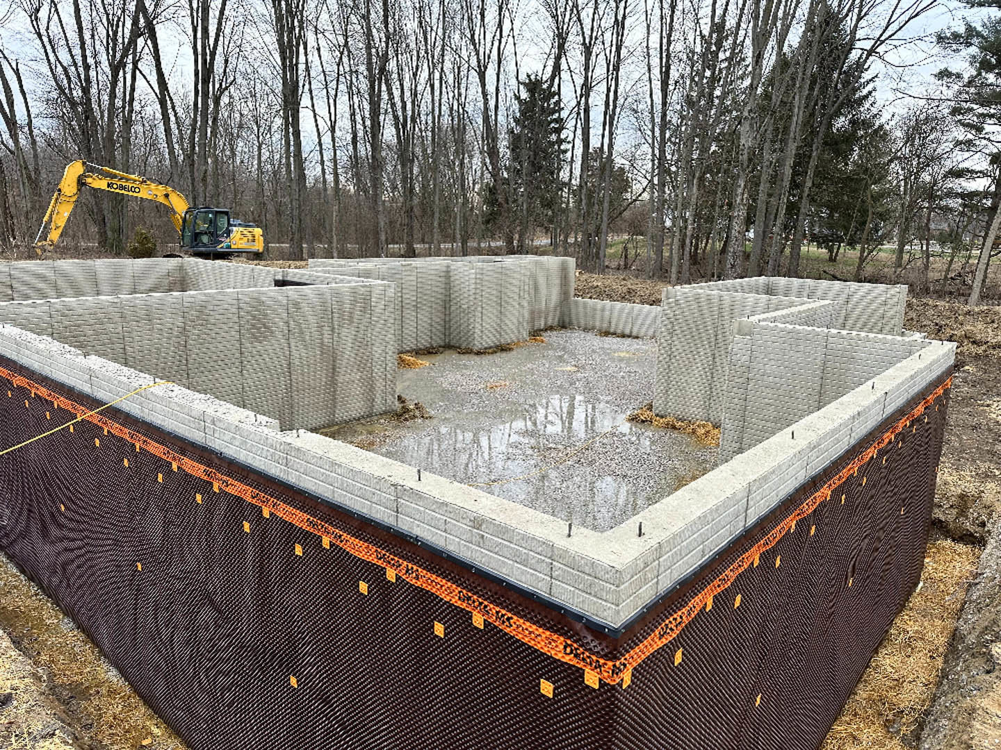 Concrete foundation covered with black tarp, yellow excavator parked beside wooded area, construction materials scattered around large custom home site.