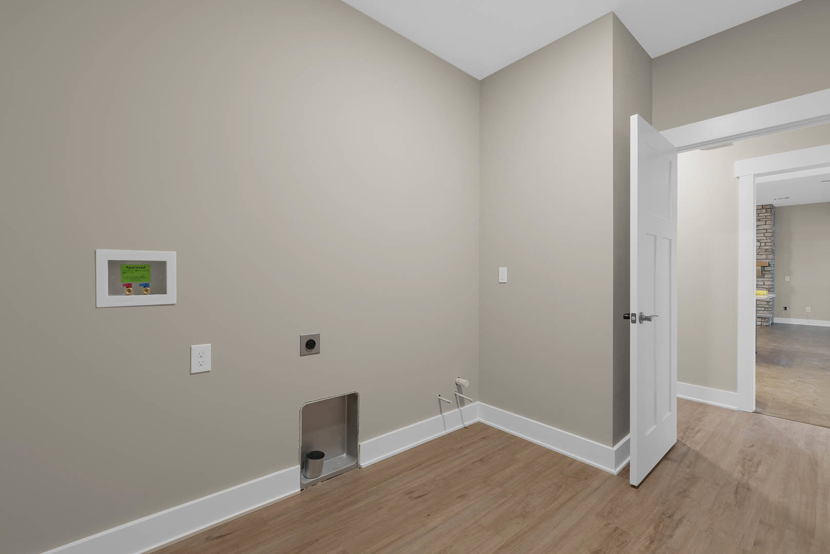 Open door leading into a room with wood flooring and white baseboards, white plaster walls, silver trash can on a shelf, green sign on a white surface, and a white double wall