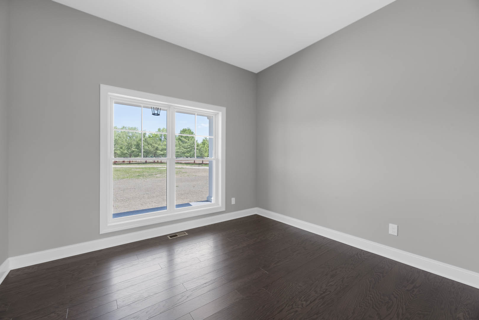 Sunlit room featuring dark hardwood flooring, white plaster walls, and a large window overlooking a field and trees.