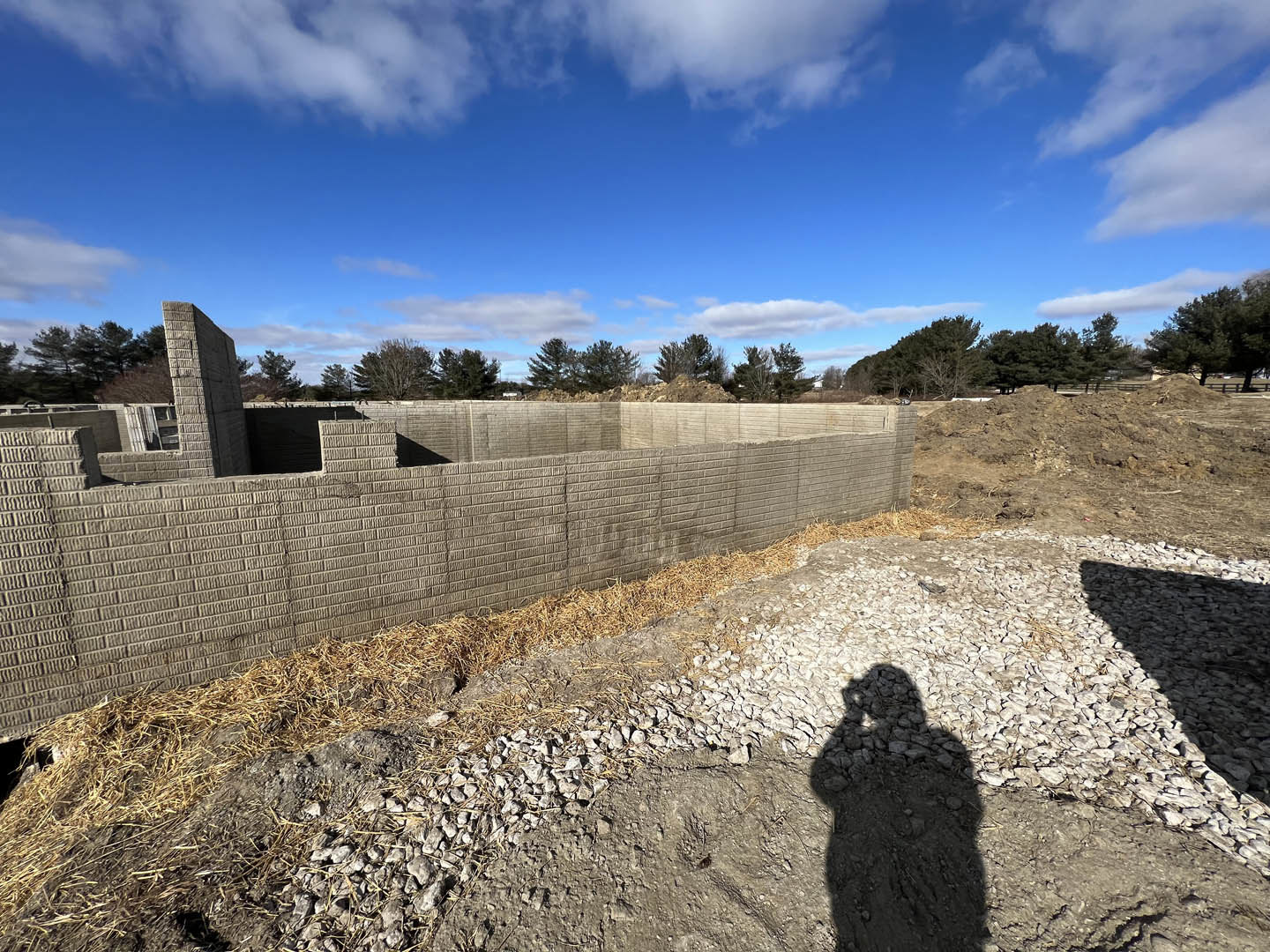 Concrete and stone exterior wall with straw accents, dirt path and rocky ground in foreground, blue sky with scattered clouds, shadow of person visible on ground.
