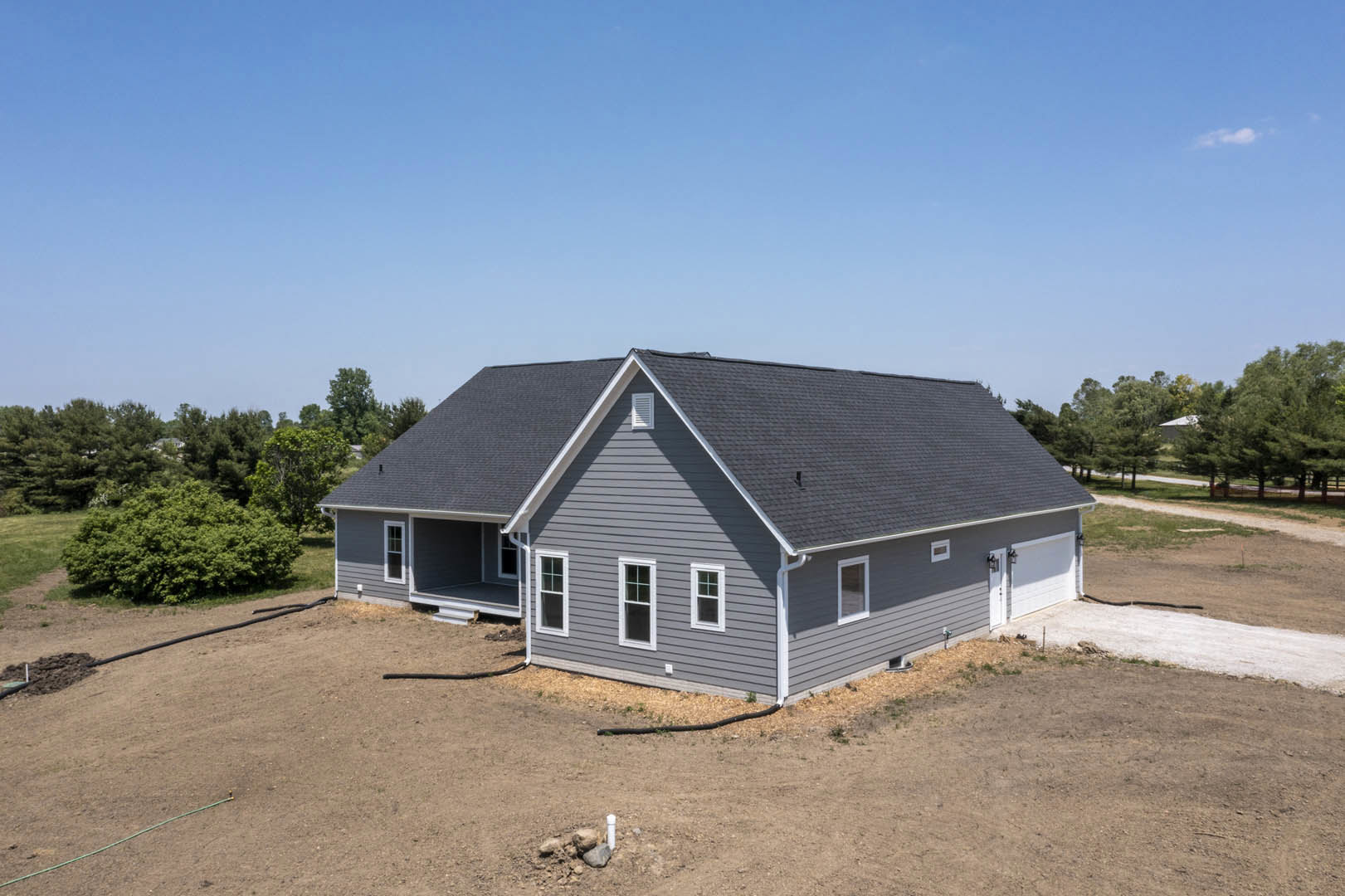 Two-story home with gray shingle roof, attached garage, paved driveway, white-framed windows, hose on side wall, landscaped front yard with tree, blue sky overhead