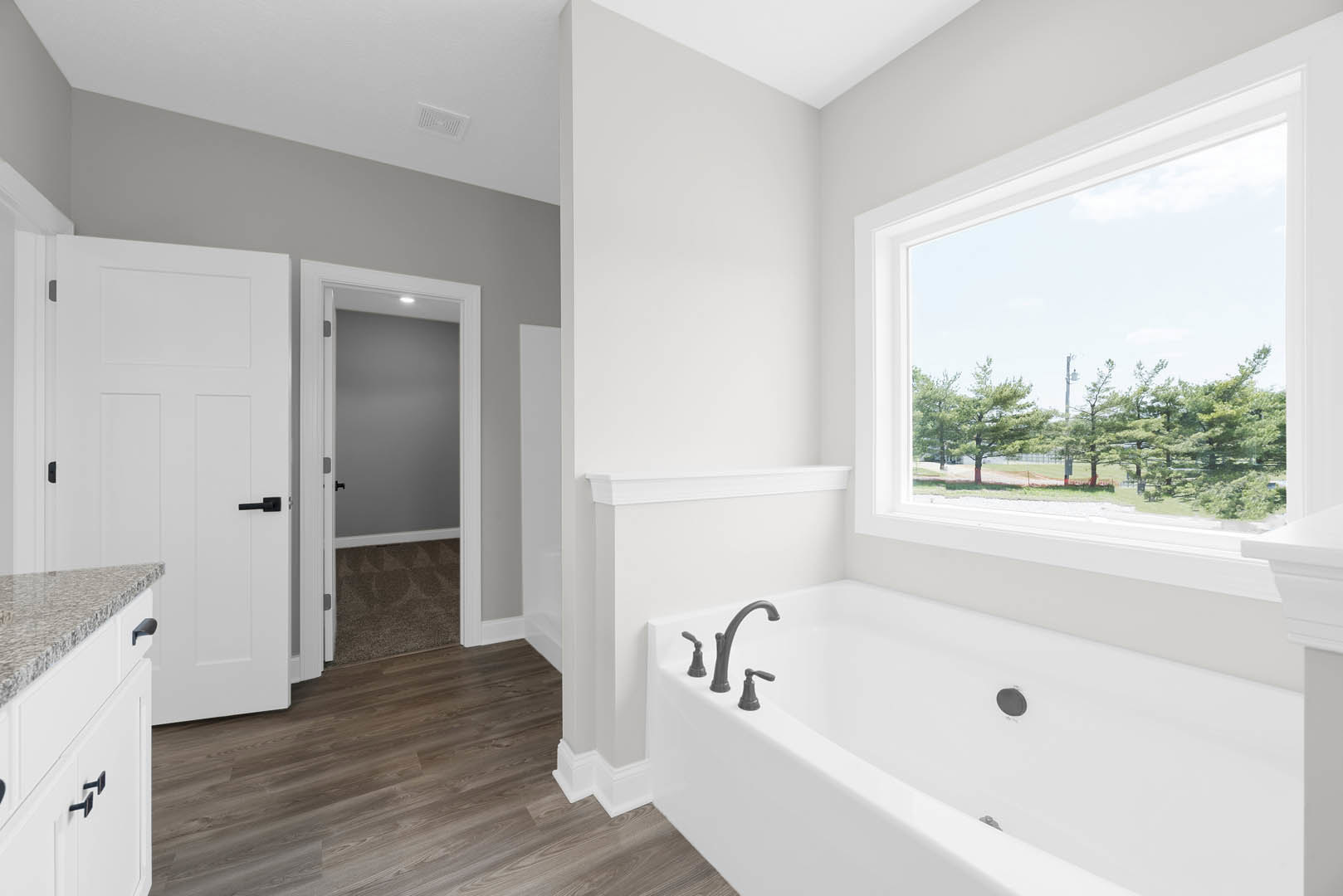 White freestanding bathtub beneath a large window with leafy trees visible outside, chrome faucet, white walls, and a white door with a black handle