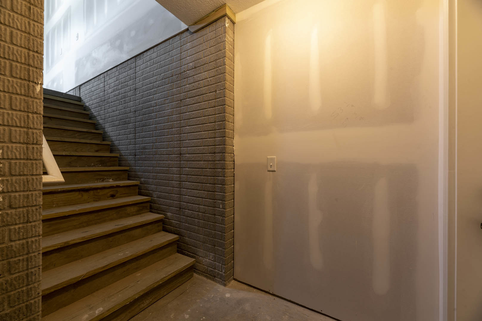 Wooden staircase with natural finish beside a painted wall featuring a light switch, exposed brick accent, and doorway in a modern interior.