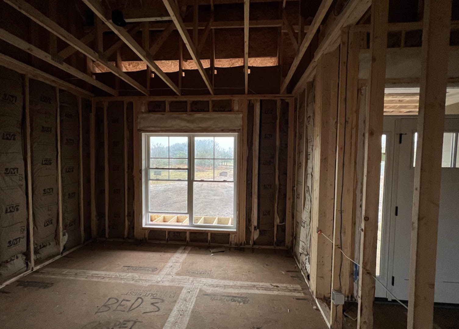 Bedroom with large window framed in wood, concrete floor, wooden shelf beneath window, bed with white linens, door leading to adjacent space, view of trees outside.