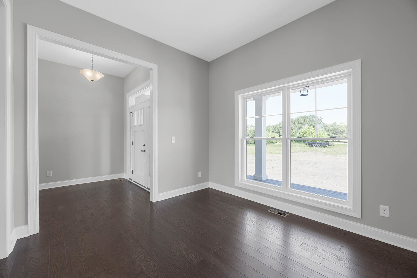 Bright room with dark wood flooring, large window overlooking lawn and picnic table, white door with black handle, ceiling light fixture, plaster walls.