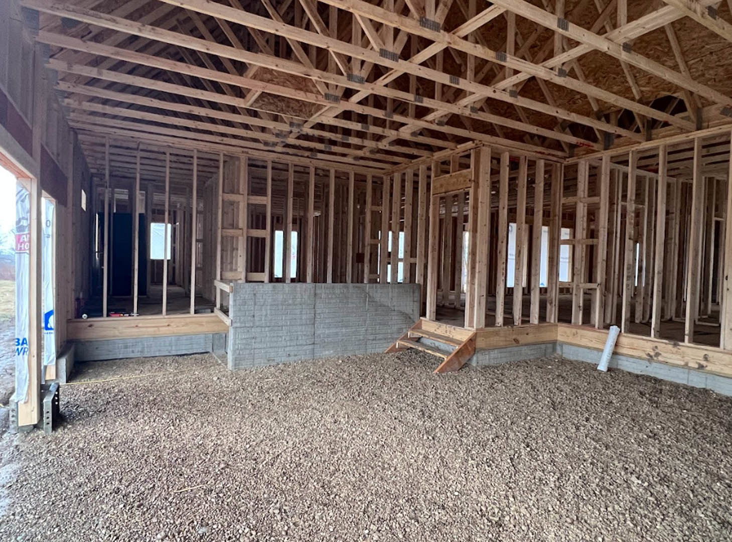 Wood-framed room with exposed ceiling beams, brick flooring, unfinished walls, and construction materials scattered on the ground