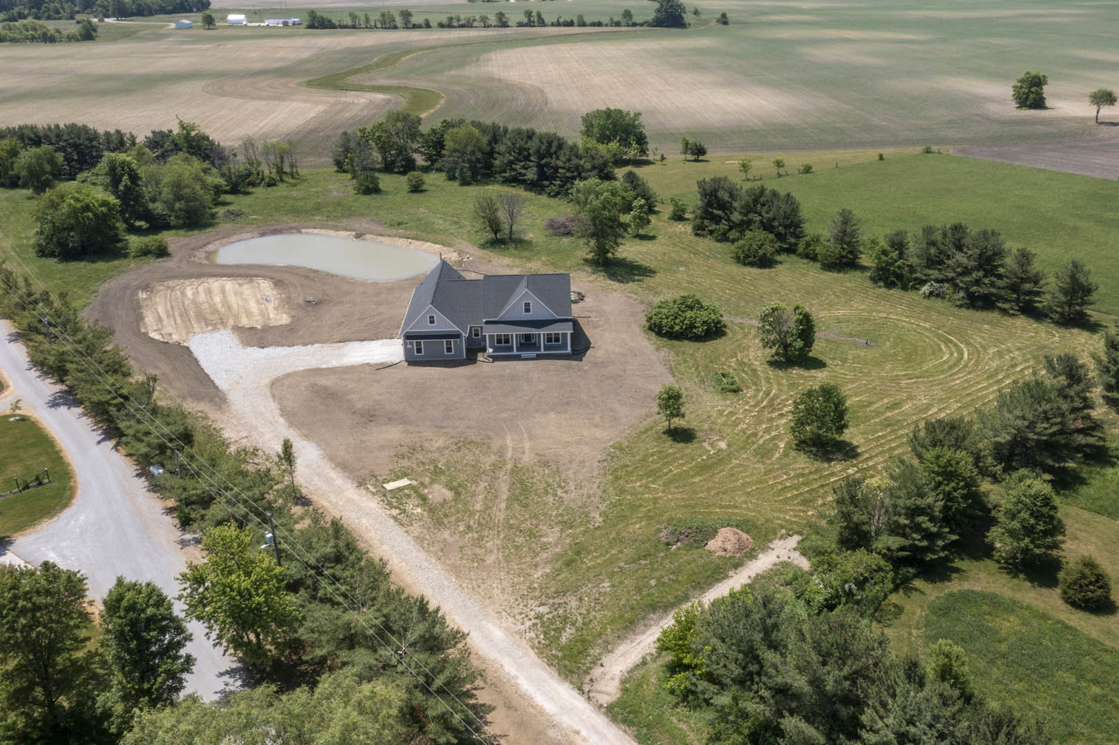White custom home with covered front porch, surrounded by grassy field, dirt mound in foreground, pond with bushes and tree nearby, rural landscape.