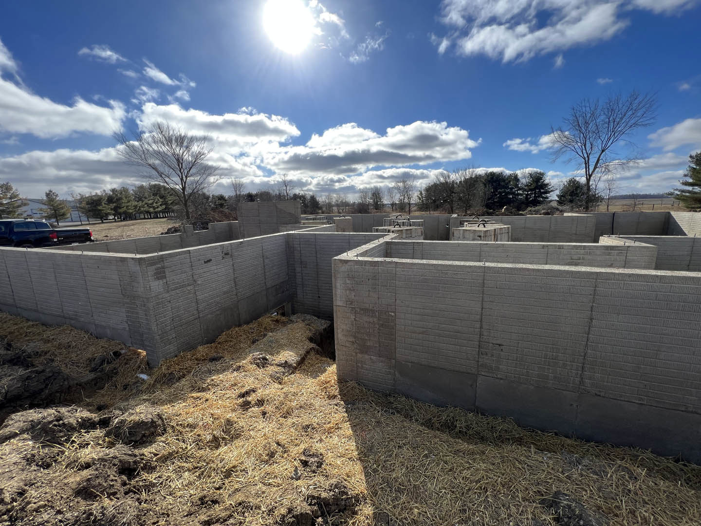 Concrete block walls and straw scattered on dirt ground, several people working at the building site, leafless tree nearby, blue sky with clouds and sunlight overhead