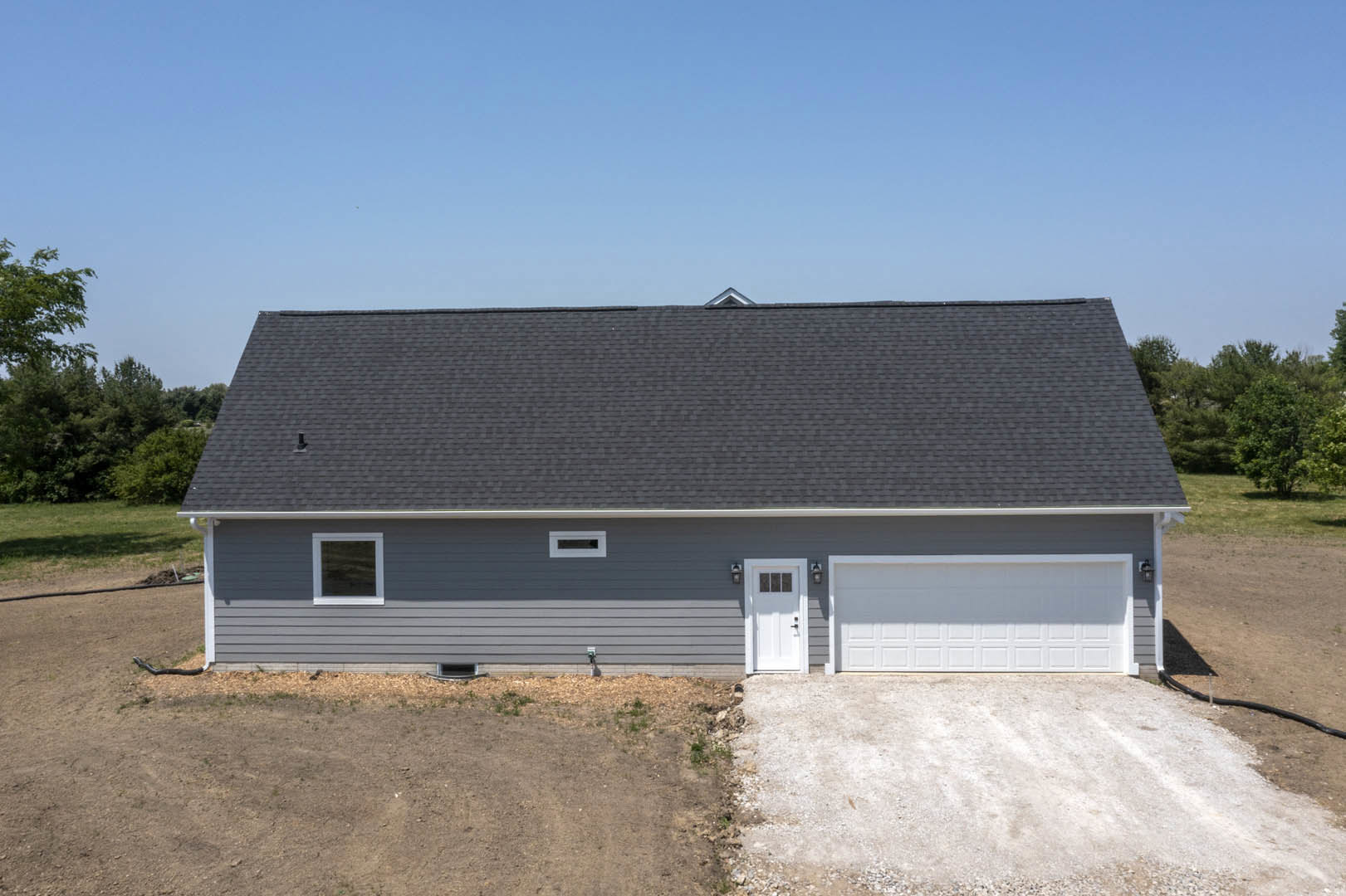Grey siding house with white-framed windows, white garage door, black shingled roof, concrete driveway, and blue sky overhead