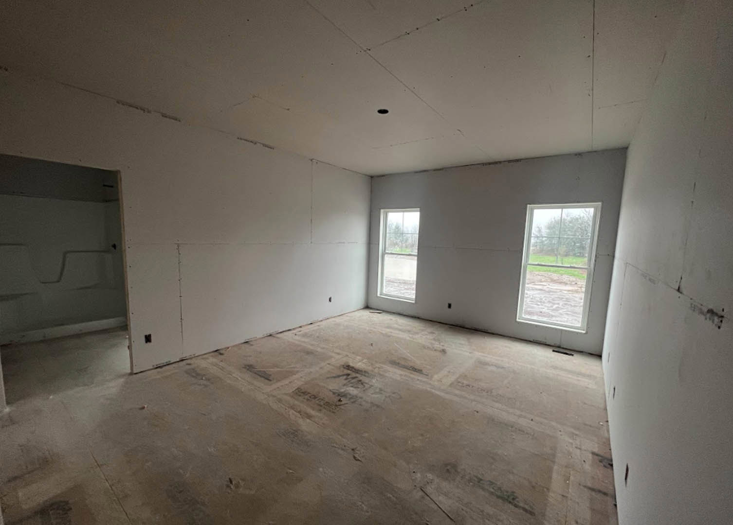 Spacious room featuring two large windows overlooking a grassy field, concrete flooring with black markings, white plaster walls, and ceiling.