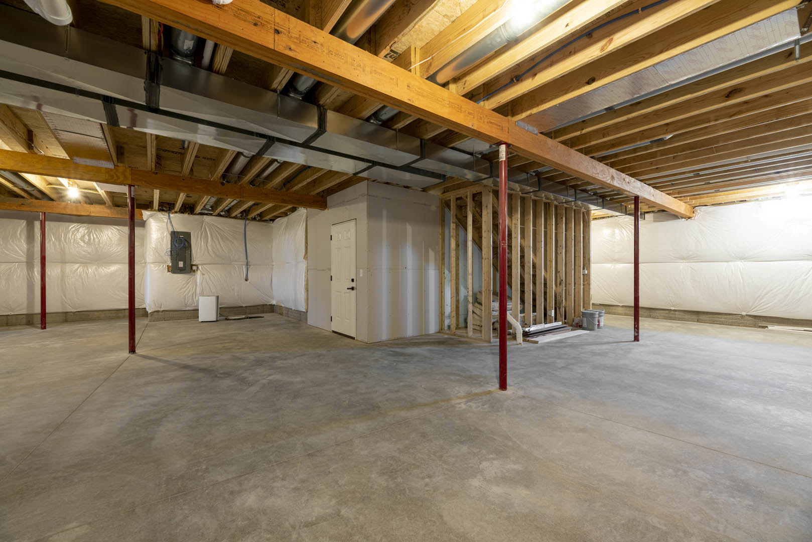Concrete floor with red support pole, exposed wood beams and metal pipes overhead, white door with black handle, white rectangular object on wood surface, unfinished basement