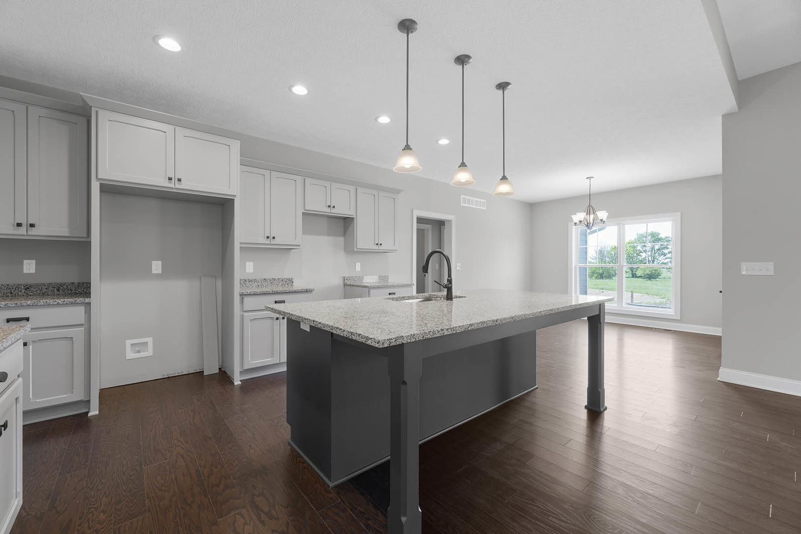 Open kitchen with wood flooring, central island featuring built-in sink and black faucet, white cabinetry, large window overlooking green lawn, white walls, and modern appliances.