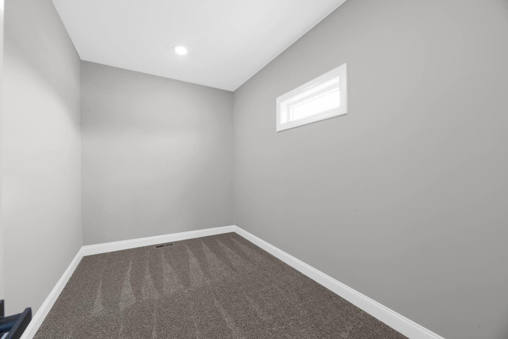 Neutral-toned carpeted floor, white rectangular window set in smooth plaster wall, ceiling light fixture, soft natural daylight illuminating simple indoor room