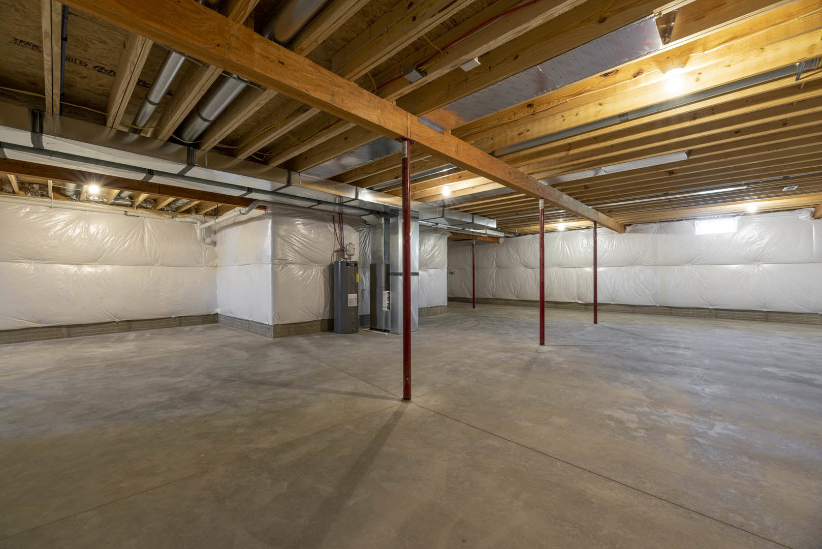 Basement room with exposed wooden ceiling beams, concrete floor, red metal support pole, and building insulation along the walls.