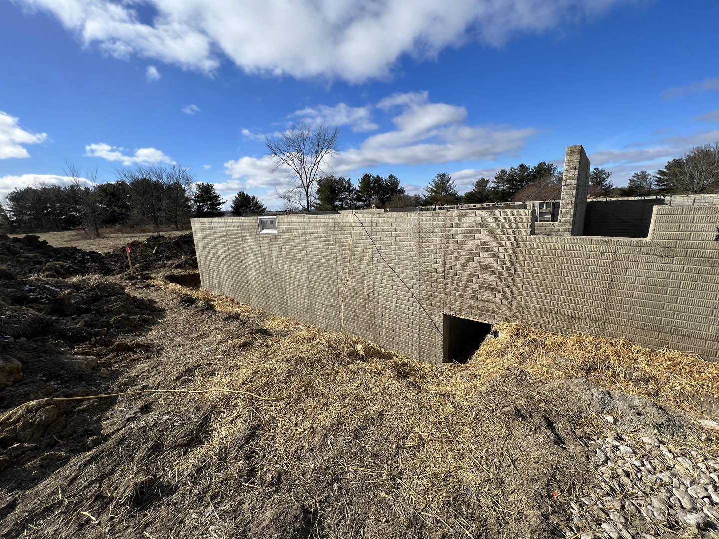 Red brick wall with a large hole, stone pillar featuring a square pattern, leafless tree, blue sky with scattered clouds