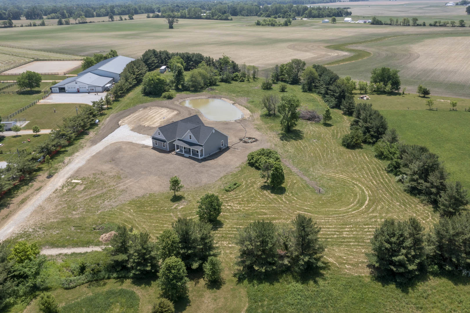 Two-story house with large covered porch, gray siding, and gabled roof, set in grassy field with scattered trees and small cement-edged pond nearby