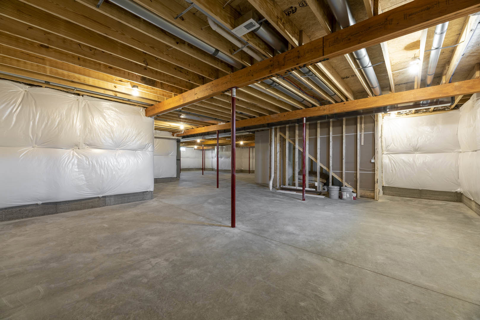 Basement room with exposed wooden beams, white plastic coverings on walls, concrete floor with red support poles, visible ceiling pipes, and large white bag against wall.