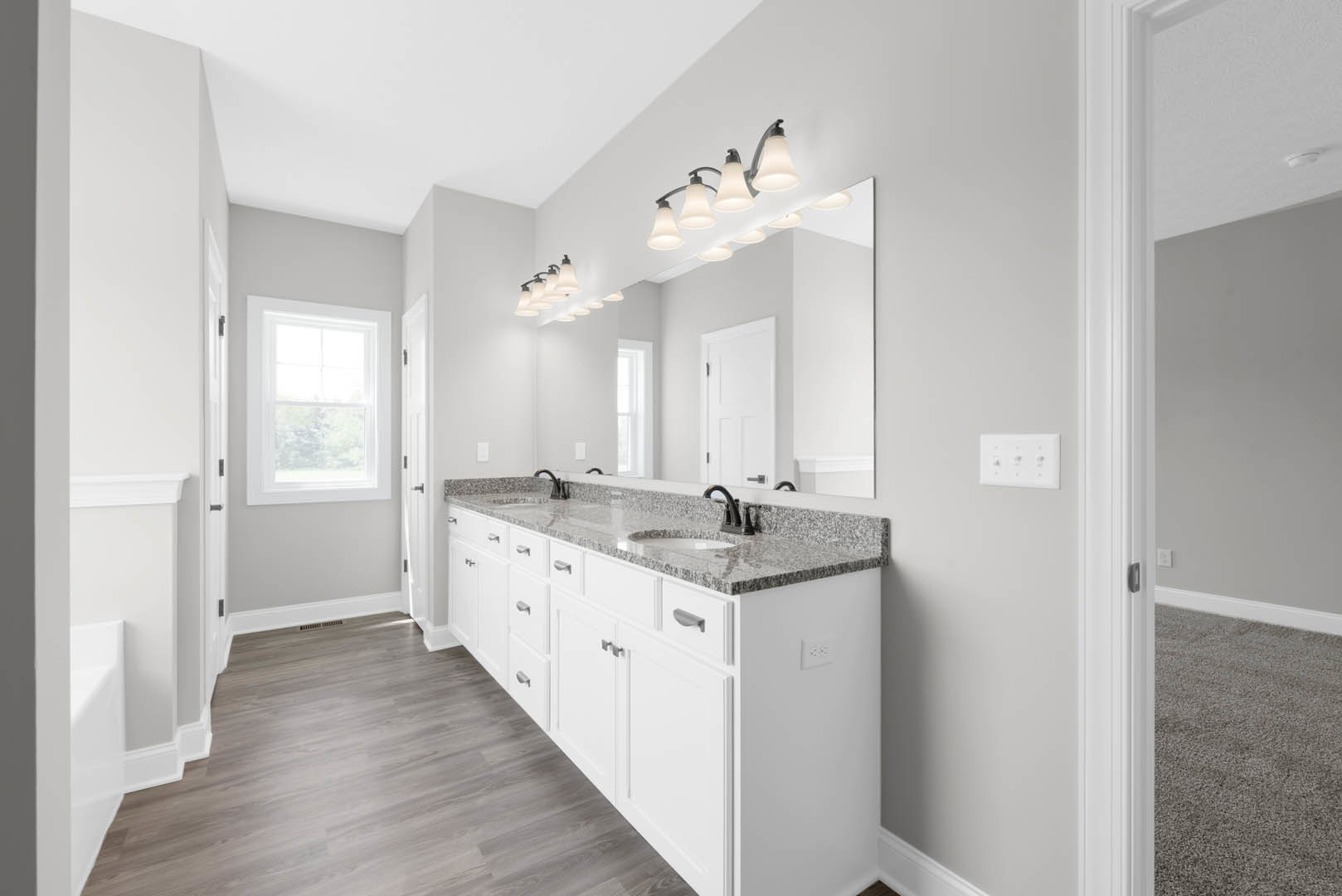 Bathroom with double vanity and wide mirror, white framed window, light wood cabinetry, tile backsplash, quartz countertop, and row of wall-mounted lights