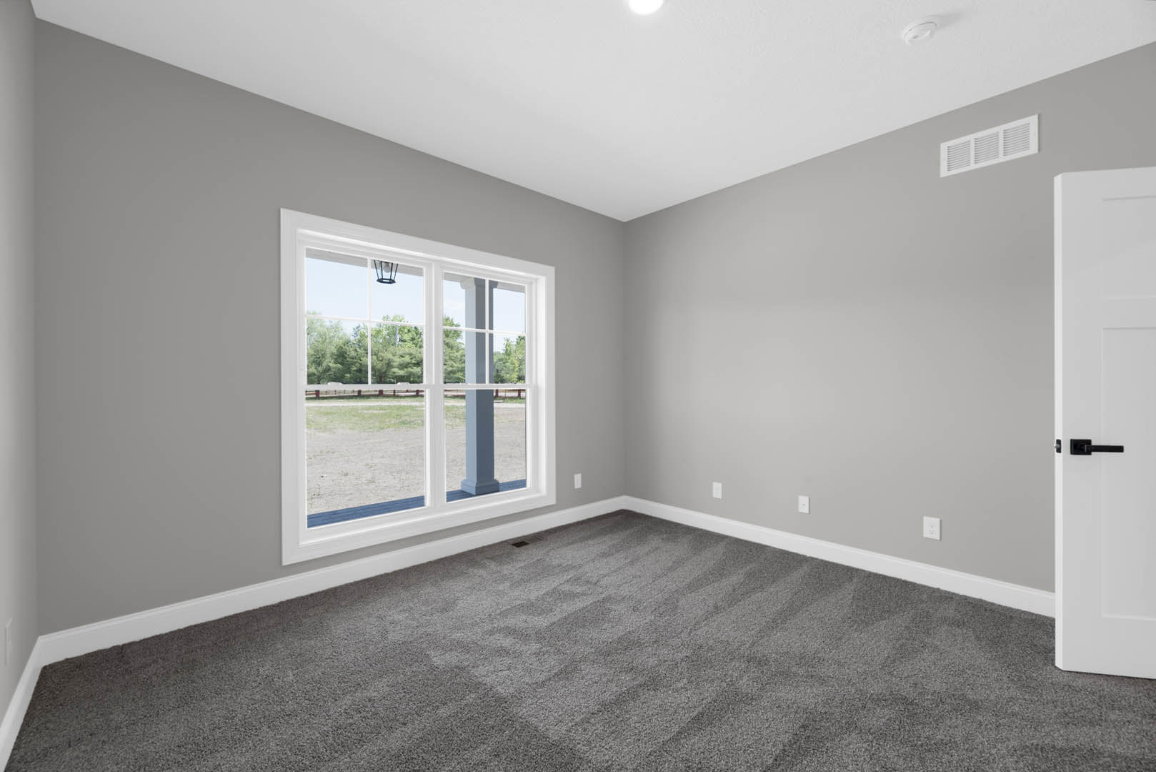 Bright room with large window overlooking grassy field, soft grey carpet flooring, white walls, modern door handle, white air vent, ceiling light fixture