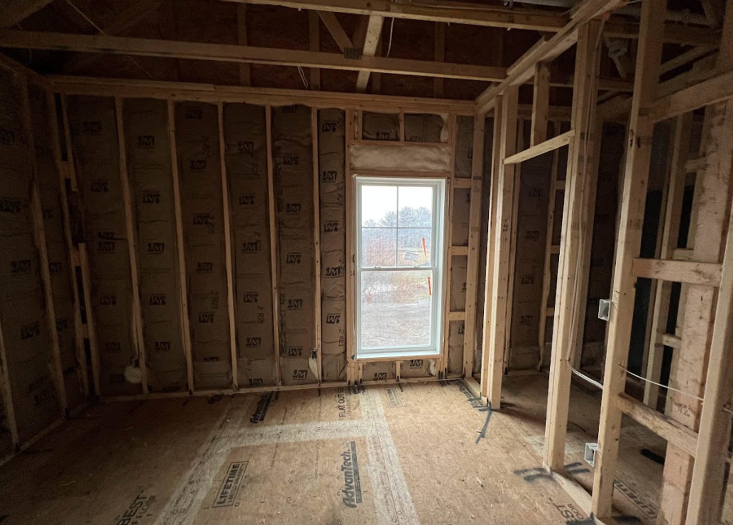 Room under construction with exposed wood framing, large window overlooking snowy landscape, wooden floor, and black floor grate