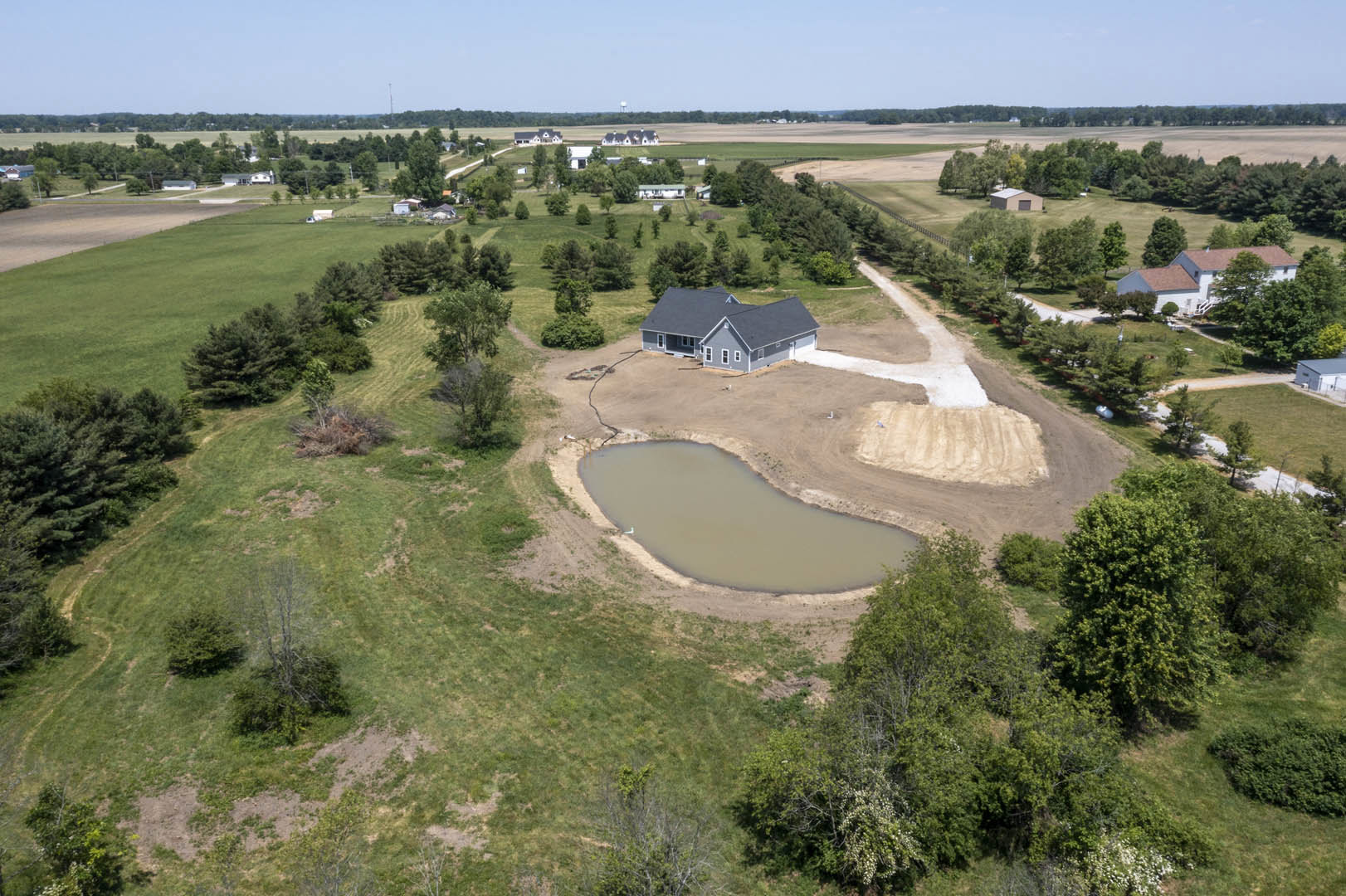 Modern house with grey roof and white walls, surrounded by a pond and landscaped grass, set against a blue sky with scattered clouds, aerial view showing trees and open field.