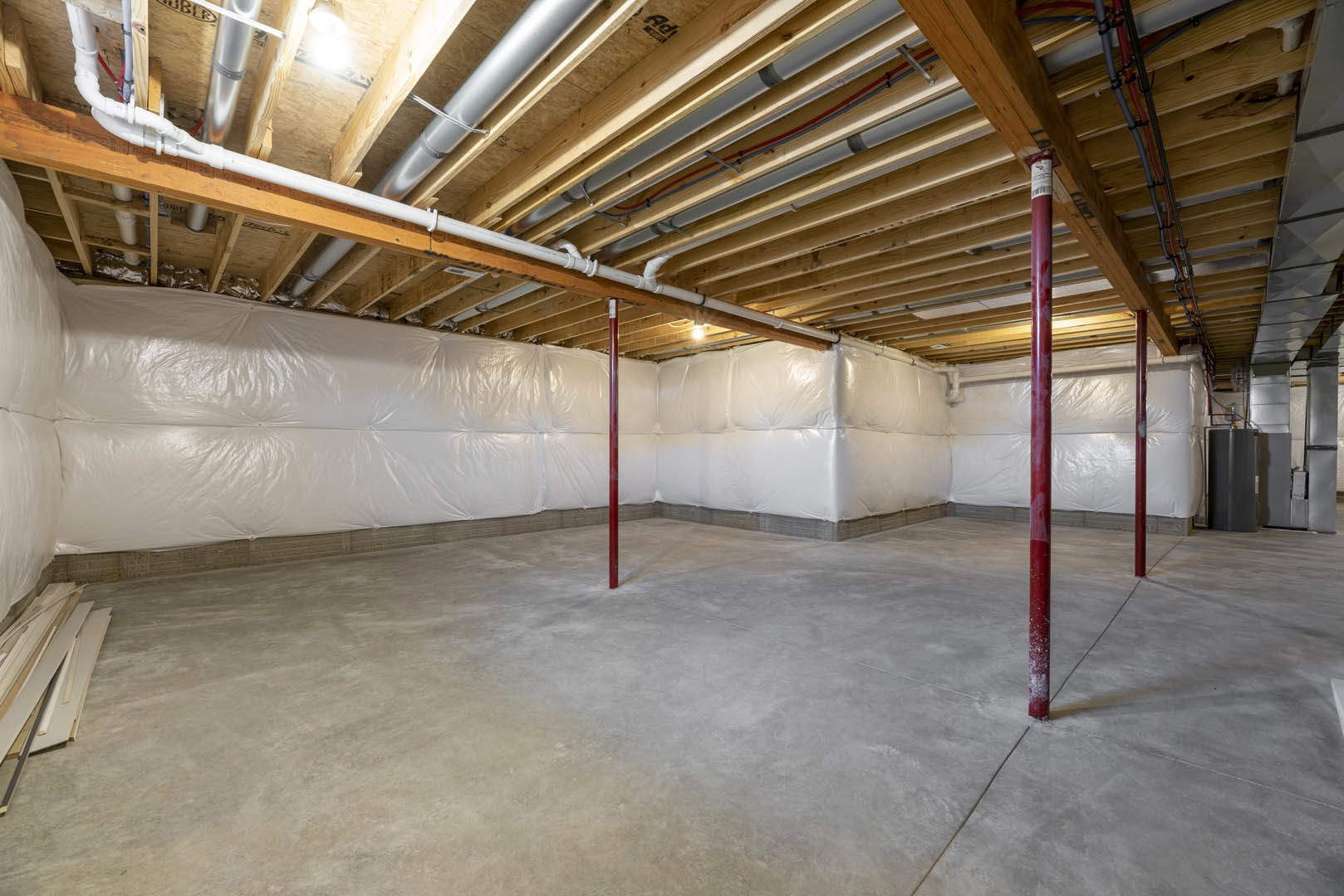 Basement under construction with exposed wooden ceiling beams, white plastic wall coverings, concrete floor, and red support pole