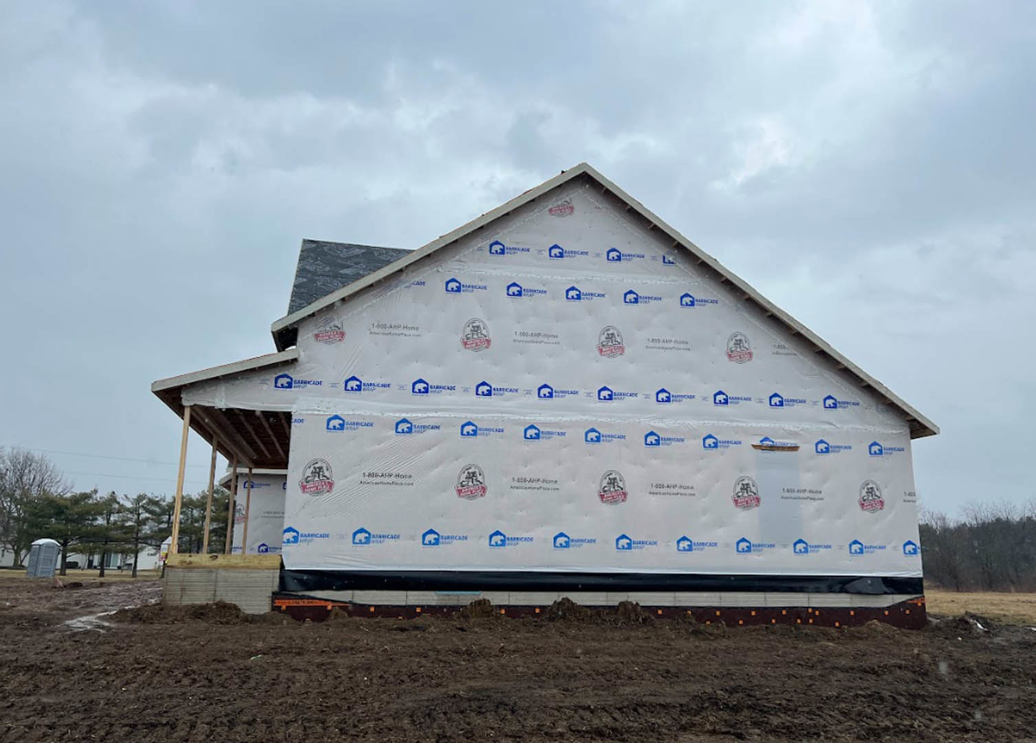 Partially built house with roof covered in white insulation, exposed framing, portable toilet in foreground, cloudy sky overhead, trees and construction materials surrounding