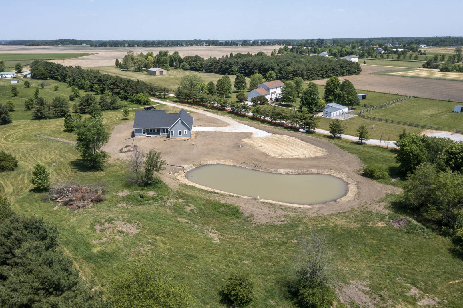 Grey-roofed house beside a pond in a grassy field, surrounded by trees under a blue sky with clouds, aerial view