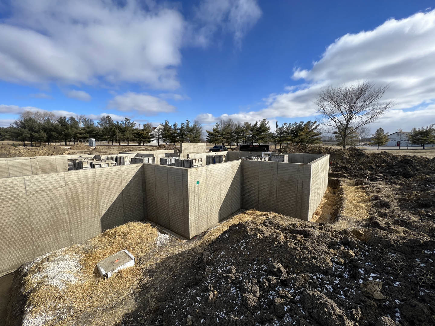 Construction site with a large dirt pile, leafless tree, concrete block in grassy area, partially built concrete wall, hay scattered on ground, and blue sky with clouds